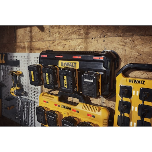 A DEWALT battery charging station mounted on a wooden wall, with several DEWALT 20V batteries inserted. Various DEWALT tools and accessories are visible nearby, including a drill and other chargers.