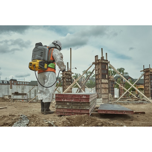 A person wearing protective clothing and a helmet uses a DEWALT backpack sprayer at a construction site with concrete pillars and wooden supports.