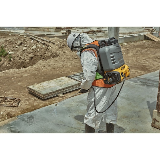 A person wearing protective clothing and a helmet is spraying water or chemicals onto a concrete surface using a DEWALT backpack sprayer, at a construction site.