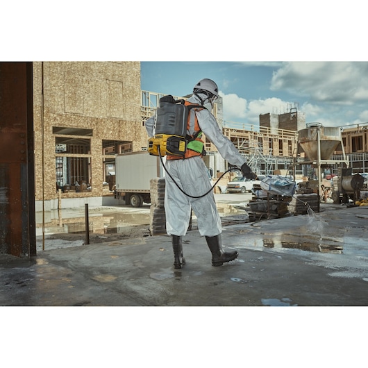 A person wearing protective clothing, boots, and a helmet uses a DEWALT backpack sprayer to spray liquid on a concrete surface at a construction site. The background shows unfinished buildings, construction materials, and equipment.