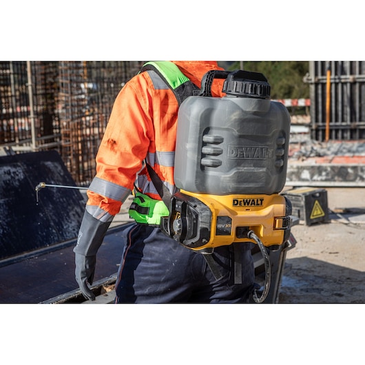 A construction worker wearing an orange high-visibility jacket and safety gloves is using a DEWALT backpack sprayer (model DCCS1100N) at a building site.
