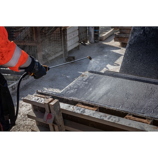 A person wearing a high-visibility orange jacket and black gloves uses a pressure washer to clean a metal surface outdoors on a construction site.