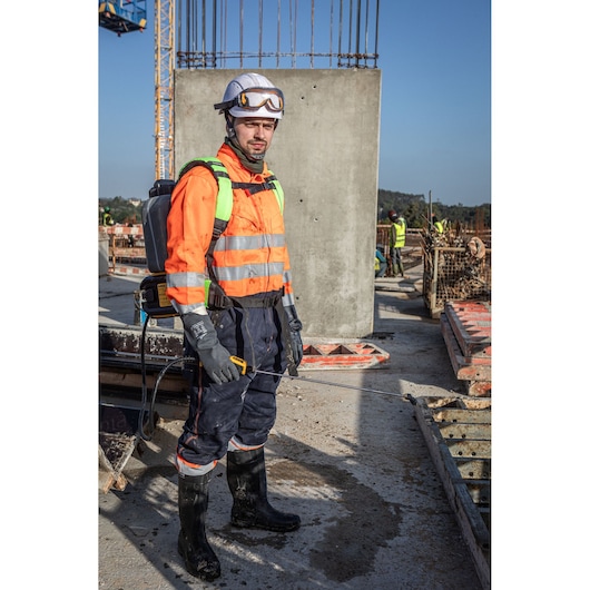 A person wearing high-visibility protective workwear, gloves, boots, and a helmet with safety goggles, standing on a construction site holding a DEWALT backpack sprayer. The individual’s face is blurred for privacy.