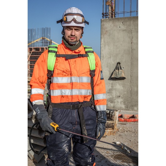 A construction worker wearing an orange safety jacket with reflective stripes, a helmet, and gloves is holding a DEWALT tool at a construction site. The worker's face is blurred, and there are concrete structures and equipment visible in the background.