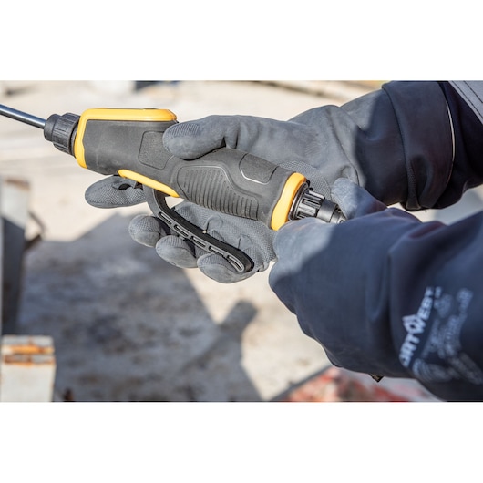 Close-up of a person wearing grey gloves and holding a DEWALT power tool with a black and yellow handle, being operated outdoors.