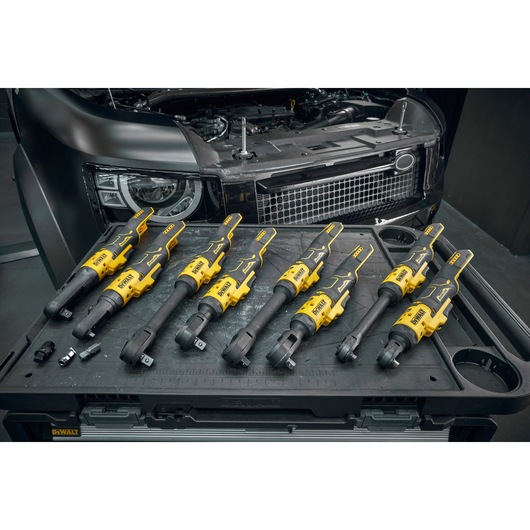 A set of Dewalt cordless ratchet tools displayed on a workstation, with a car engine and front grille visible in the background. Various tool attachments and sockets are also placed on the workstation.
