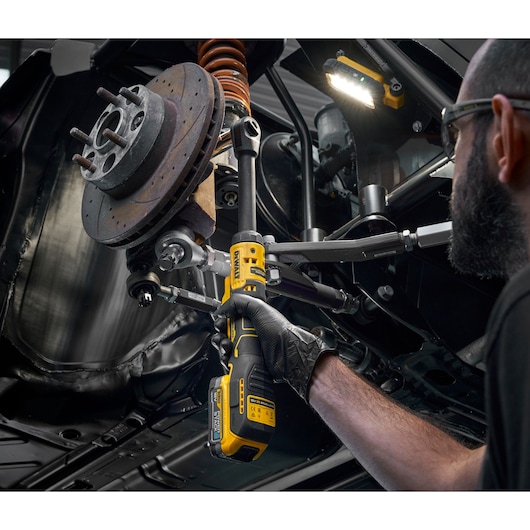A person is using a yellow and black DEWALT cordless ratchet tool on the suspension components of a vehicle. The individual is wearing black gloves and safety glasses while working underneath the car, which is elevated. There is also a DEWALT work light illuminating the area.