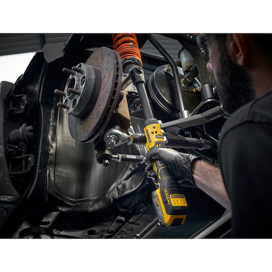 A person uses a DEWALT DCF513EN cordless ratchet tool to work on the suspension and brake components of a vehicle. The tool is yellow and black, and the person is wearing a black glove, performing automotive maintenance.