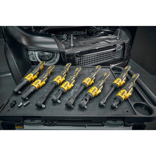 Several yellow and black DEWALT cordless ratchet wrenches are arranged on a work surface in front of a partially opened vehicle hood, with other small tool accessories also visible.