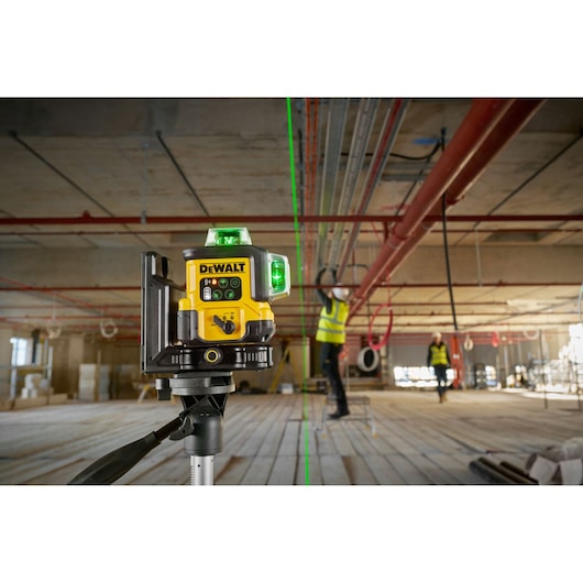 DEWALT laser level device projecting a green laser line in an indoor construction site, mounted on a tripod, with workers in safety vests working in the background.