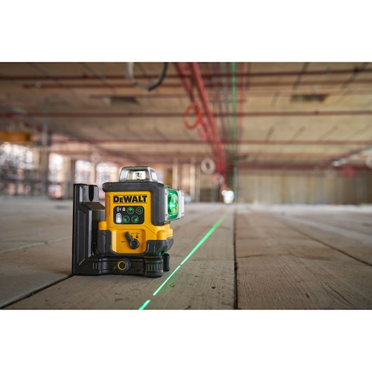 A DEWALT laser level device placed on a wooden floor in a construction site, projecting bright green horizontal and vertical laser lines across the space.