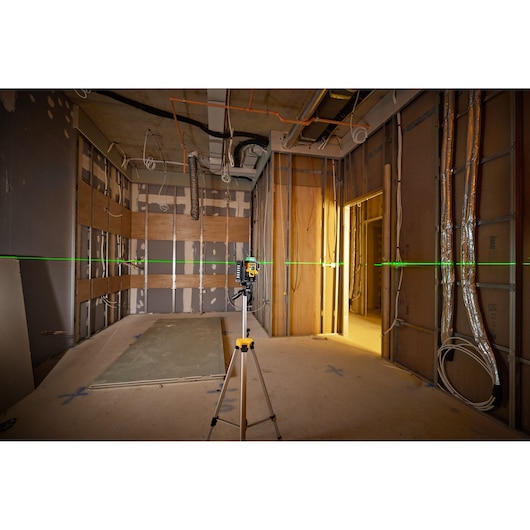 A DEWALT laser level placed on a tripod projects a green horizontal laser line across an unfinished room with exposed walls and electrical conduits.