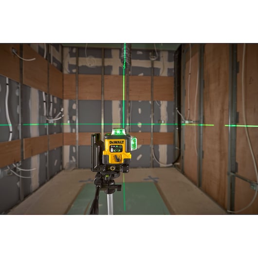 A DEWALT laser level device emits green horizontal and vertical laser lines inside a partially constructed room, mounted on a tripod. The walls show exposed wiring and partially finished surfaces.