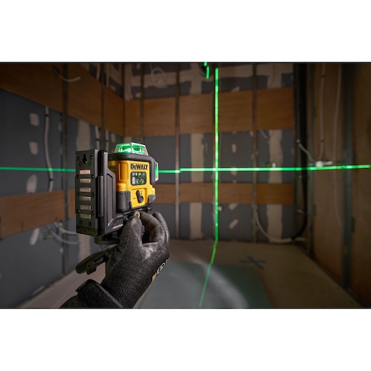 A gloved hand holds a DEWALT green laser level, model DCLE14361GB, projecting horizontal and vertical green laser lines onto the unfinished walls of a construction site.