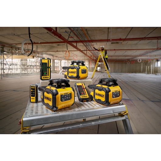 A group of DEWALT yellow and black laser levels and measuring devices are displayed on a metal platform in a large construction site. In the background, a worker in a high-visibility vest stands on a ladder with scaffolding and pipes visible throughout the unfinished building.