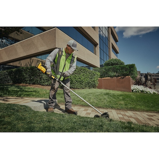 A person wearing a high-visibility vest and work gloves uses a DEWALT cordless lawn edger (model DCMED411N) to trim the edge of a grass lawn near a sidewalk outside a commercial building.