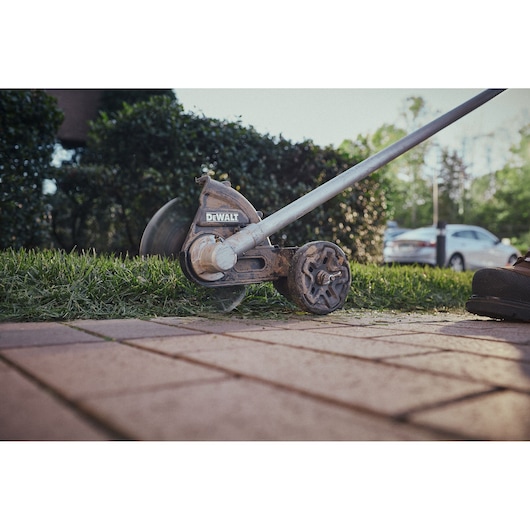 Close-up view of a DEWALT grass edger trimming the edge of a lawn next to a paved walkway, with greenery and parked cars in the background.