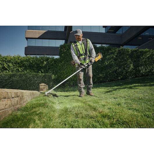 A person wearing work clothes and safety gear uses a DEWALT cordless string trimmer (SKU: DCMST901N) to trim grass along the edge of a lawn next to a brick wall, with a building and hedges in the background.