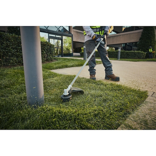 A person is using a DEWALT cordless string trimmer to cut grass along the edge of a sidewalk in a landscaped area near a commercial building.