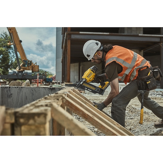 Worker uses DEWALT DCN910 nailer on wood at construction site, wearing safety helmet and orange vest.
