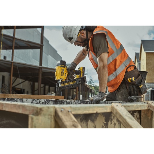 Worker using DEWALT DCN910 nailer on construction site, wearing safety gear and tool belt.