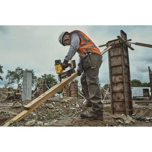 Worker using DEWALT DCN910 nailer on wood at a construction site, wearing safety gear and orange vest.