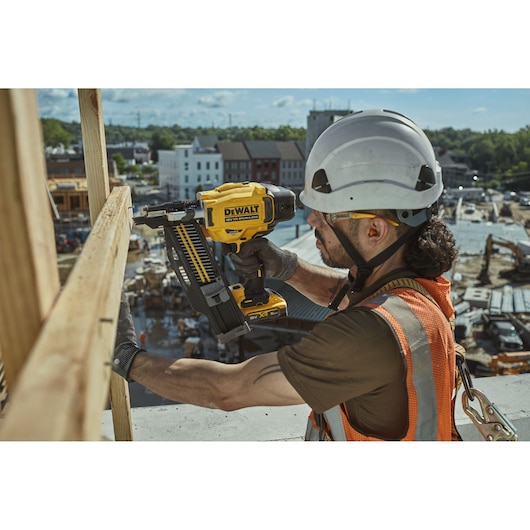 Worker uses DEWALT DCN910 nailer tool at a construction site, wearing safety gear and helmet.