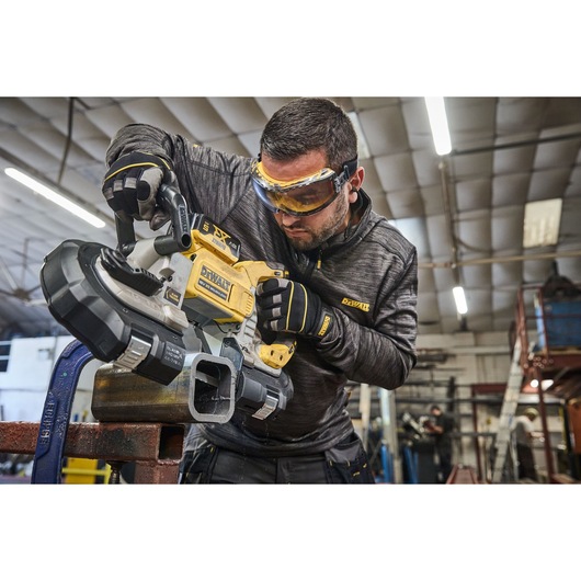 Person using a DEWALT DCS374N portable band saw to cut a metal pipe in an industrial workshop. The individual is wearing protective gloves and a dark jacket with DEWALT branding.