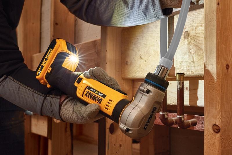 A person wearing work gloves uses a DEWALT DCE400D2 18V cordless pipe crimping tool to connect a pipe inside a wooden framed construction area.